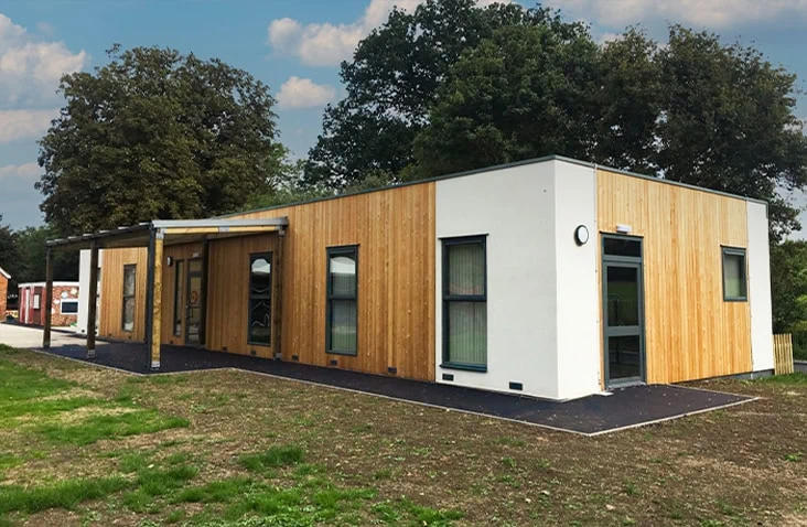 Modern modular school building with timber cladding and white panel sections, featuring large windows and a covered entrance, set on a landscaped site with trees in the background.