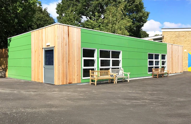 Modern modular school building with green exterior panels and timber cladding, featuring large windows and outdoor benches on a tarmac playground