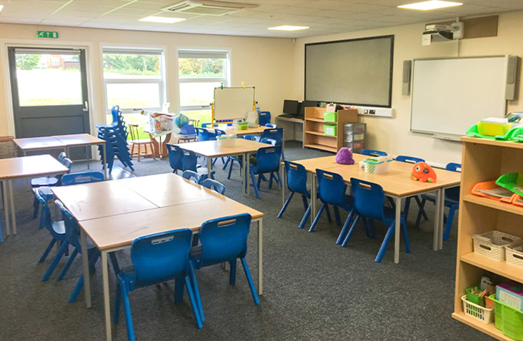 Modern modular primary school classroom with tables and blue chairs, carpeted floor, large windows, whiteboard, and organised shelving with educational materials