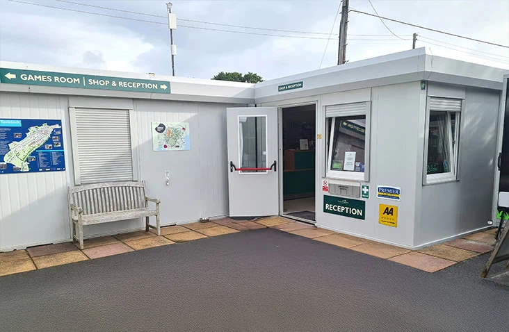 Modular reception building with grey panelled exterior, open entrance door, signage for games room and shop, and maps displayed on the wall outside.