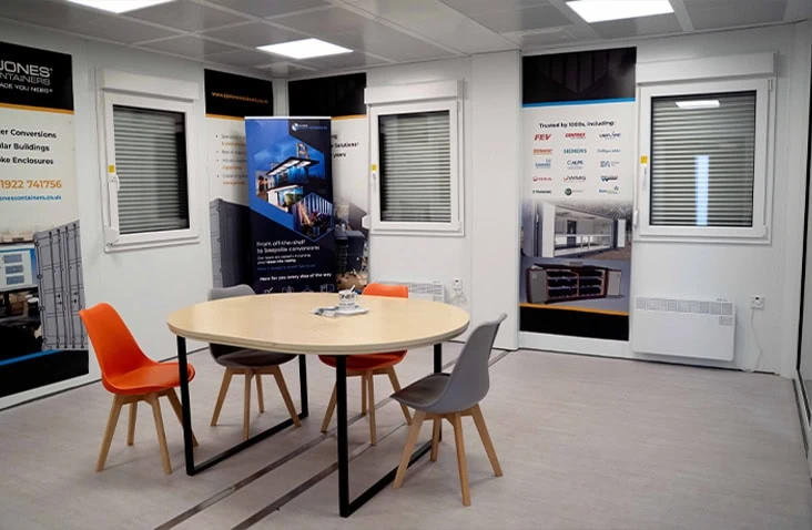 Modular office interior featuring a round meeting table with colourful chairs, wall banners displaying S Jones Containers branding, and windows with shutters.