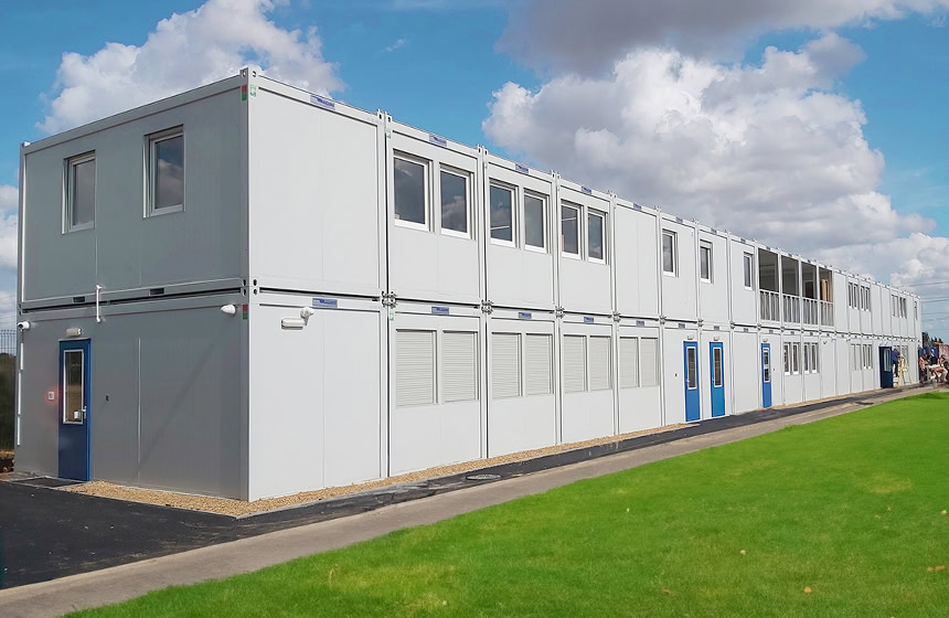 Two-storey modular school building with white panel exterior, multiple windows and blue doors, installed alongside a grass field.