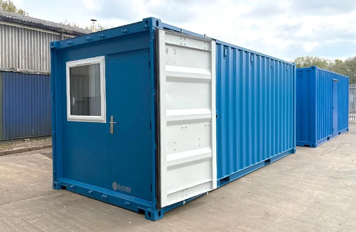 20ft blue converted shipping container workshop with a personnel door and window fitted at the front of the container revealed by open cargo doors, positioned on a concrete yard next to another similar container.