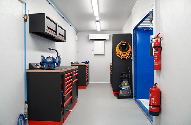 Interior of a containerised workshop fitted with workbenches, tool storage units, a bench grinder, overhead cabinets, a wall-mounted hose reel, an air conditioning unit, and fire extinguishers mounted near the entrance.