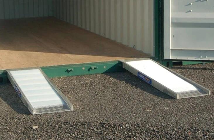 High incline container ramps positioned at the entrance of a shipping container, showing two metal ramps placed on gravel to allow wheeled access into a container with a plywood floor. 
