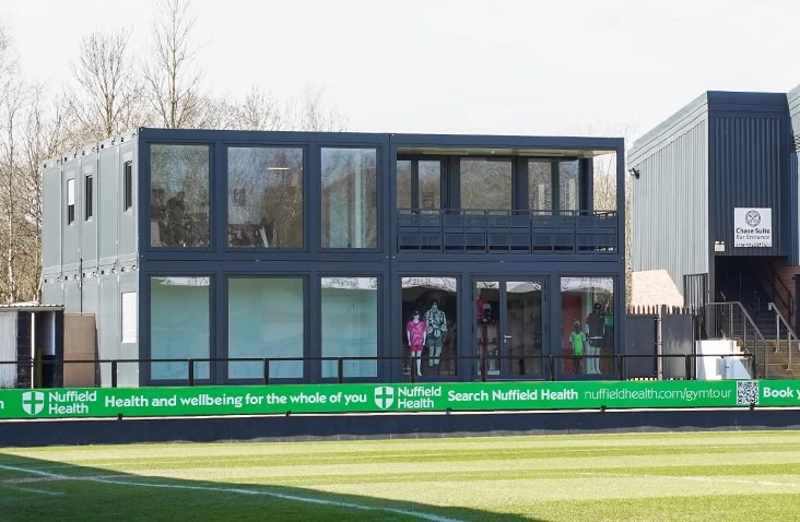 A two-storey modular building constructed from converted shipping containers at a football stadium, featuring large glass panels and balconies overlooking the pitch.