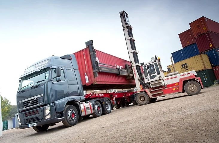 Large forklift truck loading a red shipping container onto a flatbed trailer attached to a Volvo lorry in a busy container yard.
