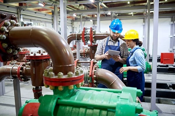 Two water treatment engineers wearing hard hats are inspecting pipework and machinery inside a water treatment facility, representing utilities and critical infrastructure. 