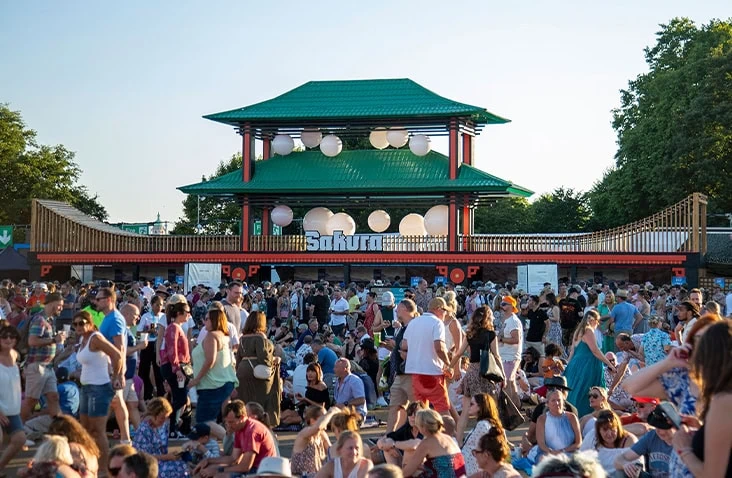 Crowded outdoor festival bar built from stacked shipping containers, featuring a green pagoda-style roof, timber cladding and Sakura branding, used as a large-scale containerised hospitality venue.