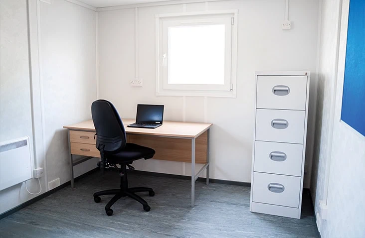 Internal office space inside a site accommodation unit, showing a desk with chair, laptop workstation, filing cabinet, window, and white insulated walls, suitable for temporary site office hire, construction offices, and modular workspace solutions. 