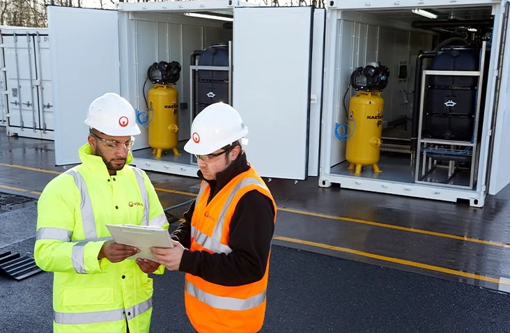 Two engineers in PPE inspecting bespoke shipping container conversions housing industrial equipment, with open containers revealing integrated plant systems and technical installations on an operational site.