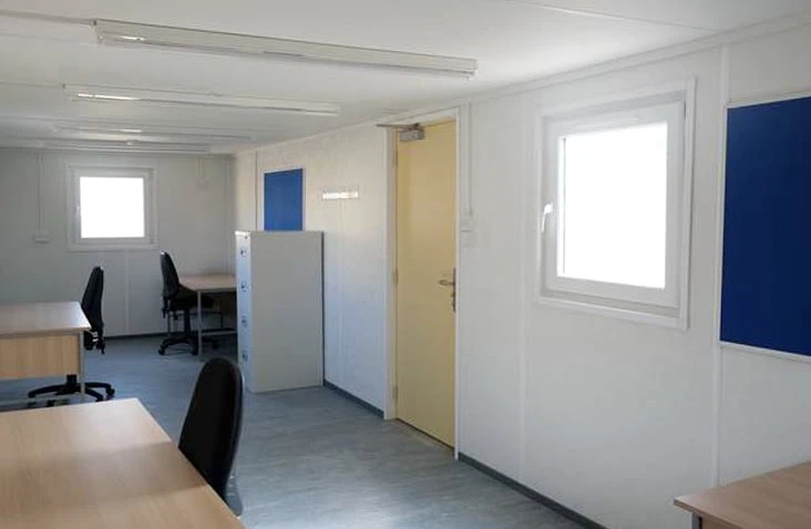     Interior of a white site office with desks, office chairs, filing cabinets and notice boards, featuring windows and fluorescent lighting, designed for temporary office accommodation hire on construction and industrial sites. 