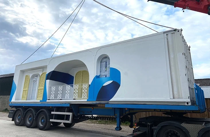 White and blue-branded shipping container being craned onto a trailer during delivery, showcasing expert transport and installation of a containerised hospitality structure for The House of Peroni.