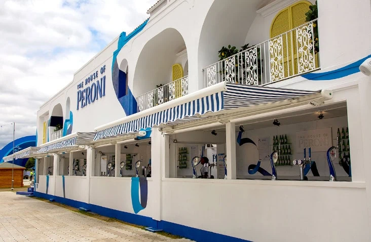 Completed container bar at The House of Peroni, formed from white shipping containers with blue detailing, open serving hatches, striped awnings and decorative balconies for a premium outdoor event venue.