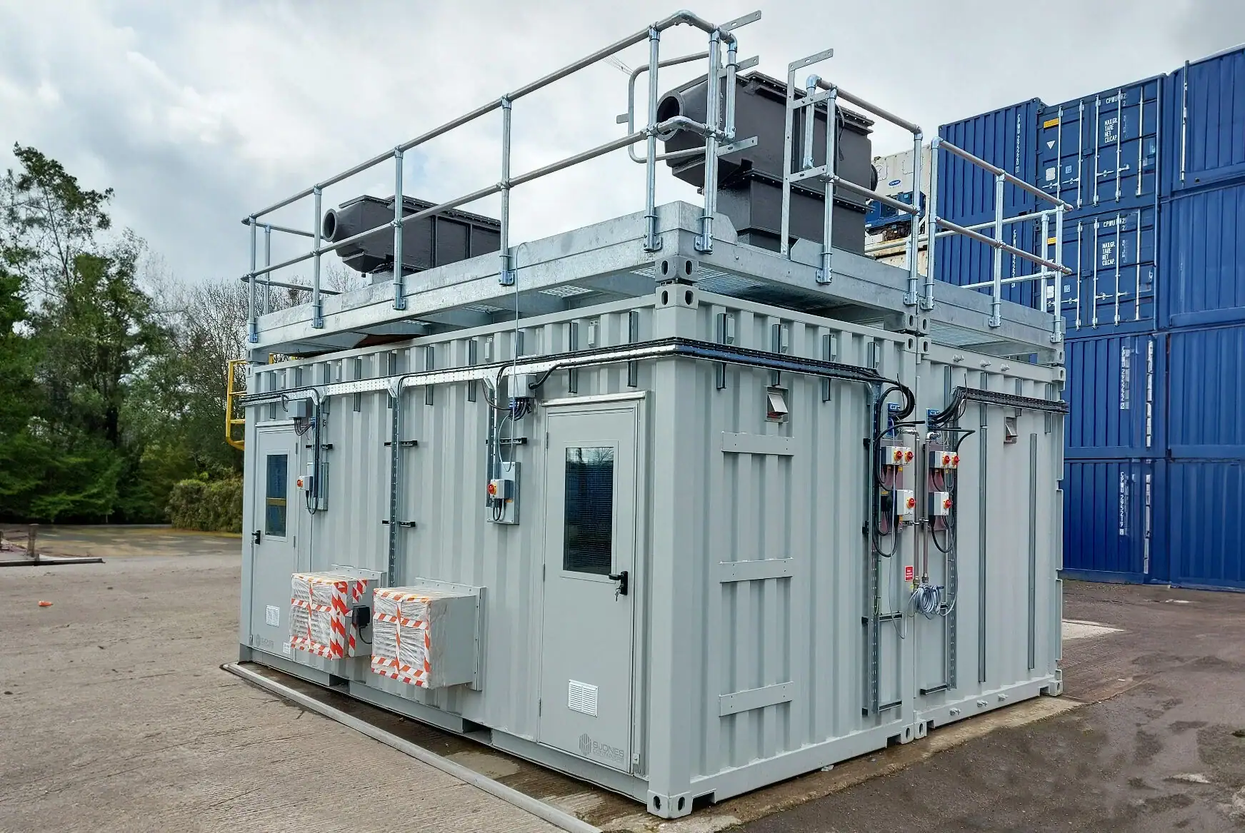Containerised battery testing unit built from a grey shipping container, featuring roof-mounted electrical equipment, external control panels, and safety enclosures, shown on an industrial yard with stacked blue containers in the background, representing battery testing and energy storage testing infrastructure.
