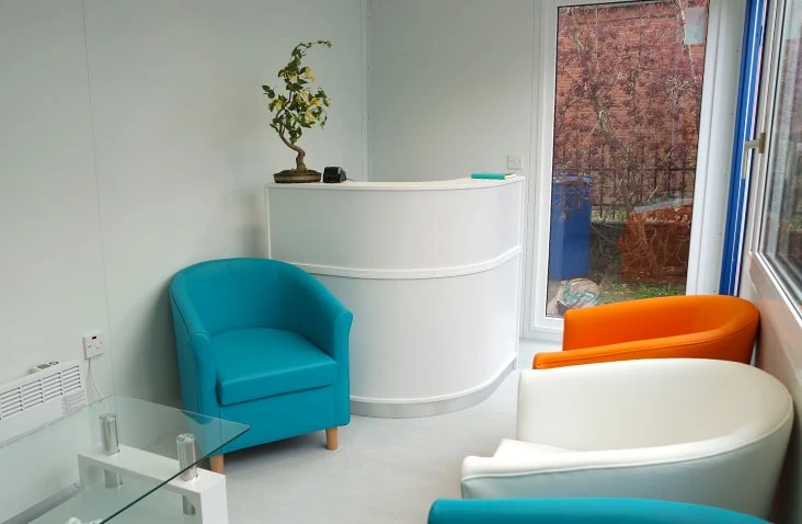 Interior of a modular reception unit featuring a curved white reception desk, colourful lounge chairs, glass coffee table and large window, designed as a modern customer waiting area for commercial or office use. 