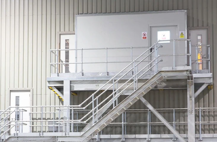 Externally mounted containerised control room conversion with steel access staircase, safety handrails and raised platform, installed inside an industrial facility to provide safe access to an elevated control and monitoring environment.