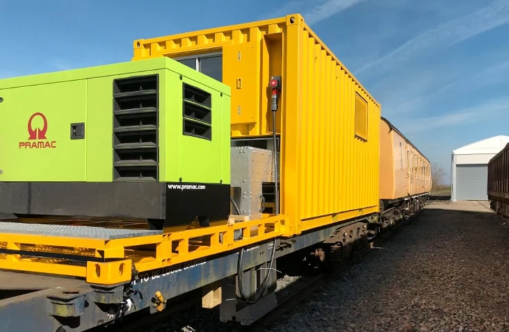Yellow containerised test unit mounted on a rail wagon and connected to a green industrial generator, demonstrating a mobile container conversion for testing, power supply and transport applications.