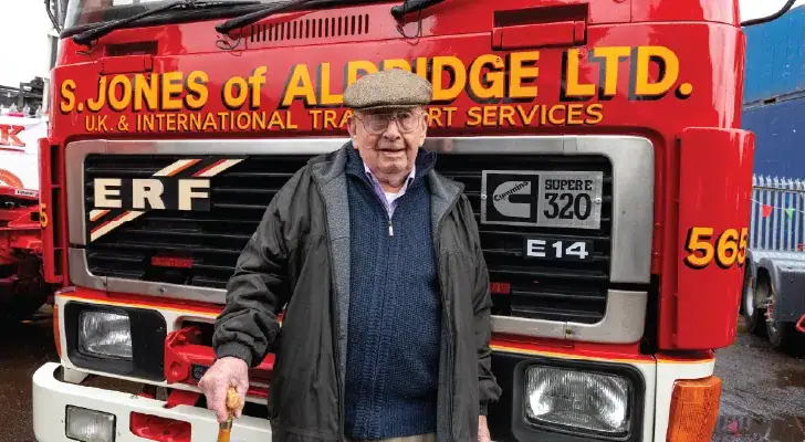 An elderly man wearing a flat cap, glasses, and a dark coat stands in front of a red ERF truck belonging to "S. Jones of Aldridge Ltd." He is holding a walking stick and has a slight smile on his face. The truck has bold yellow lettering and a Cummins "Super 320" badge.