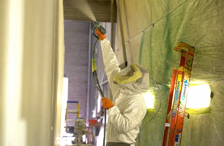 A person wearing protective coveralls, gloves, and a respirator is spraying paint onto a vertical surface inside a paint booth. A red ladder and bright work lights are visible in the background, along with green protective sheets covering the walls.