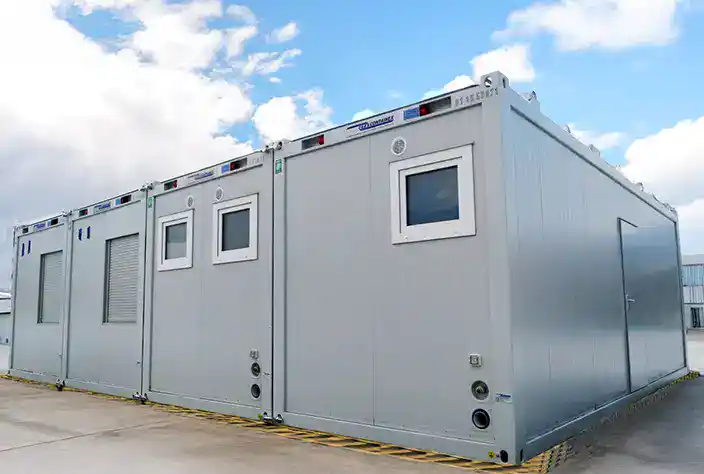 A row of modern modular container units with white panel exteriors, windows, vents, and utility connections, arranged side-by-side on a paved surface under a partly cloudy sky.