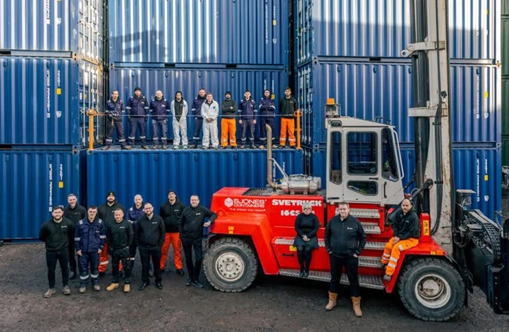 S Jones Containers team posing with a heavy-duty container lift truck in their West Midlands container depot, surrounded by stacked blue shipping containers, showcasing logistics, handling, and container conversion operations.