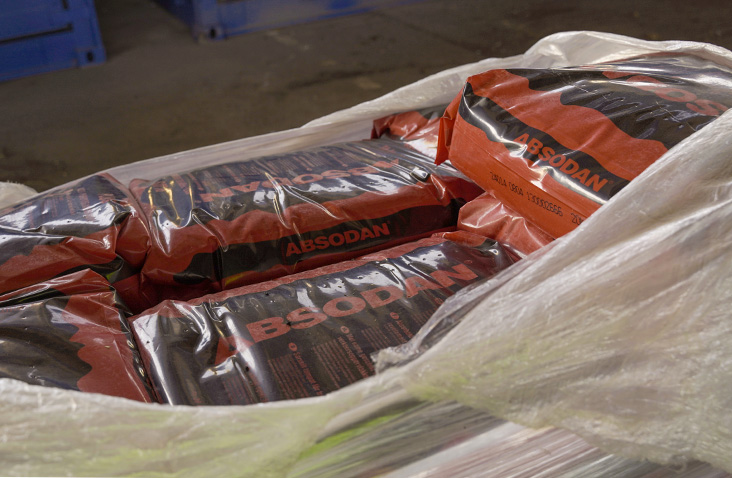 Close-up of red and black Absodan absorbent material bags stacked on a pallet and wrapped in clear plastic, inside an industrial warehouse setting.