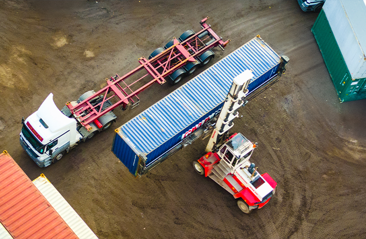Shipping container being lifted by a container handler and loaded onto a flatbed trailer at the S Jones Containers depot, ready for delivery to a customer.