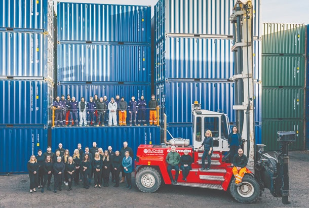 S Jones Containers team gathered at the company's Aldridge, Walsall depot, standing alongside a heavy-duty container handler and surrounded by stacked shipping containers. The group photo highlights the people behind one of the largest container depots in the West Midlands, showcasing expertise in container sales, hire, conversions, storage, handling, repairs, and logistics operations.