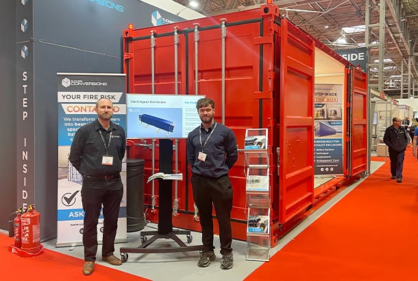 Head of Conversions Andrew Nicholls and Technical Sales Engineer Oliver Smith representing S Jones Conversions at the Fire Safety Event, standing beside a bright red 20ft side-opening shipping container conversion. The specialist enclosure showcases the company's expertise in bespoke container conversions for fire safety, switchgear, battery storage, and other engineered industrial applications.