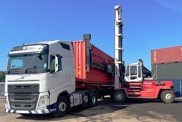 White Volvo articulated truck being loaded with a red 40ft shipping container by a reach stacker in a container yard under a clear blue sky.