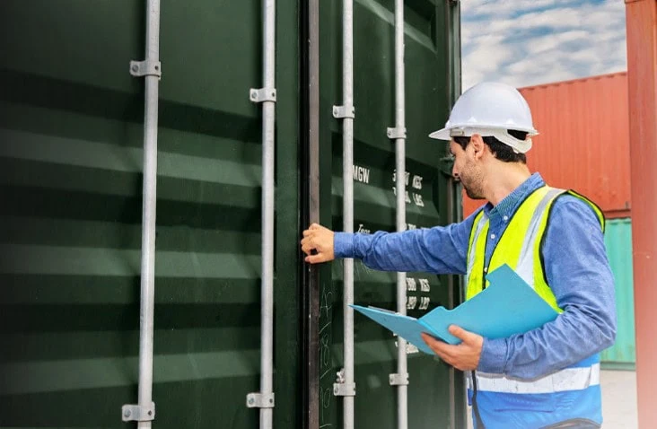 Shipping container inspection being carried out after a customer hire period, with a worker wearing a hard hat and high-visibility vest checking the doors, locking bars and exterior condition of a green steel container. Returned hire containers are inspected to identify any damage or maintenance requirements before being prepared for their next use.
